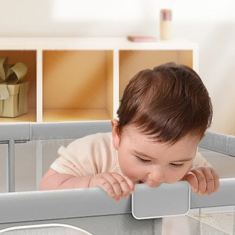 Baby leaning on gray mesh side of baby playpen gripping top rail.