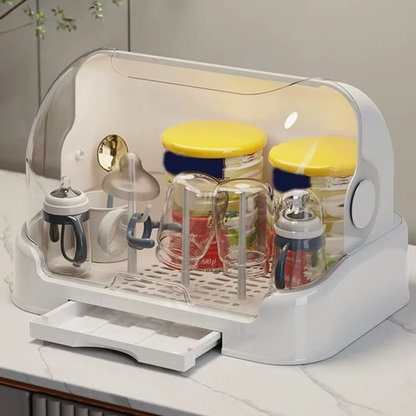 White baby bottle drying rack with open clear lid and extended drip tray on kitchen counter with bottles and jars.