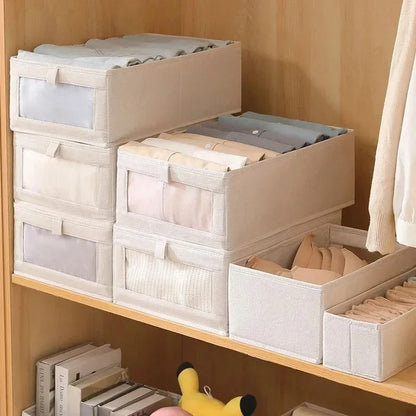 Beige foldable fabric storage boxes with clear-view fronts organized with baby clothes on wooden nursery shelves.