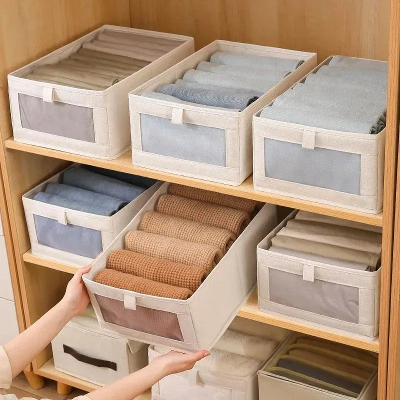 Beige foldable fabric storage boxes with clear-view front, organized with baby clothes on wooden shelves.