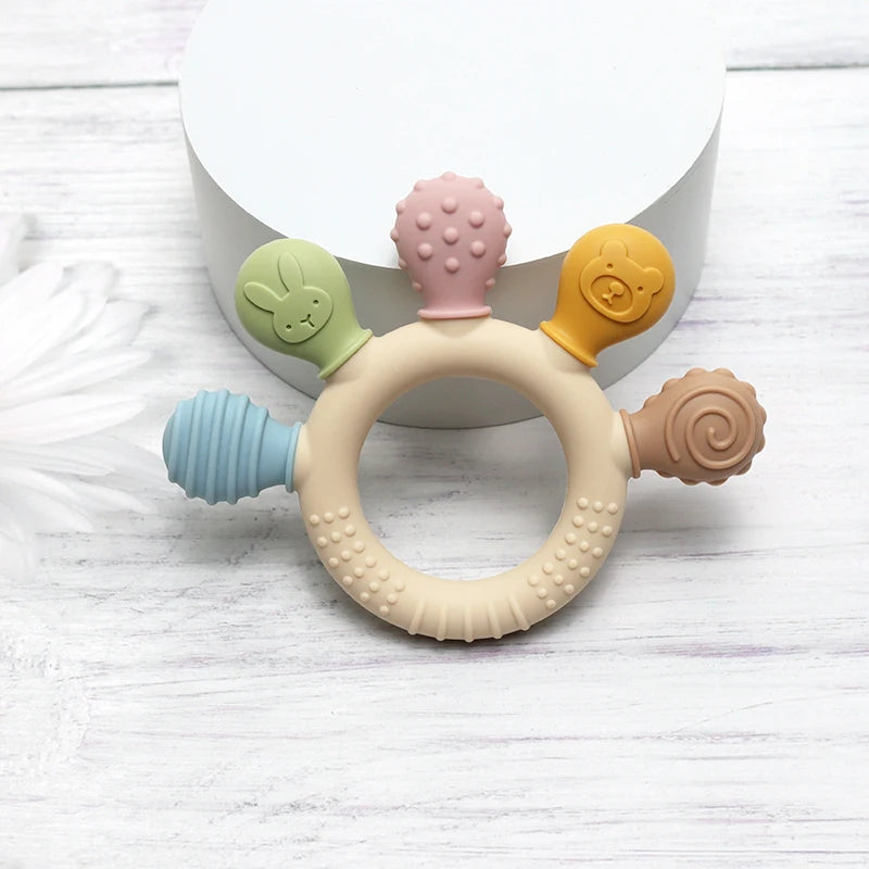 Pastel silicone baby teething ring with five textured chew heads on white wooden surface.