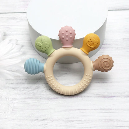Pastel silicone baby teething ring with five textured chew heads on white wooden surface.