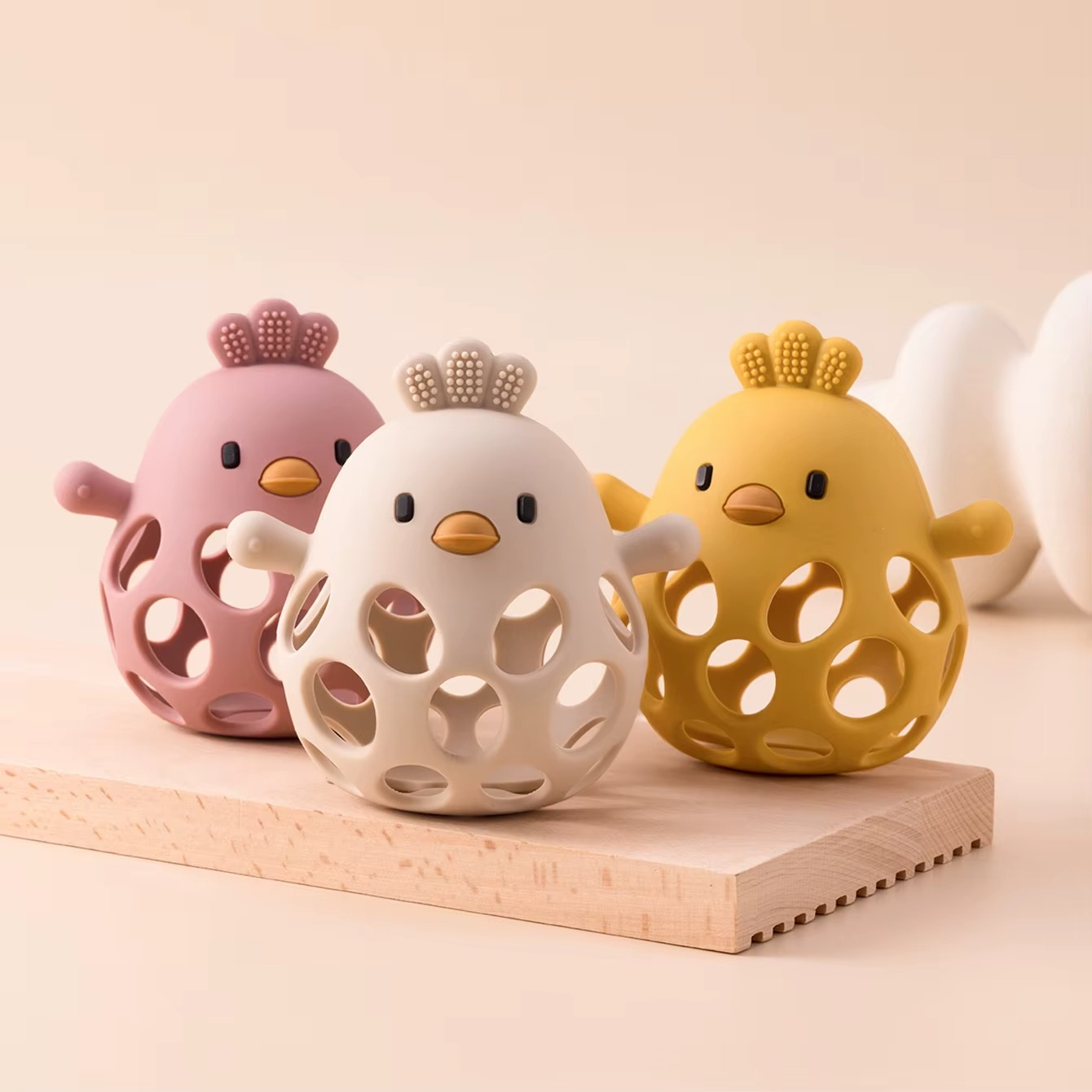 Silicone baby teether rattles in dusty pink, beige, and mango yellow on wooden board.