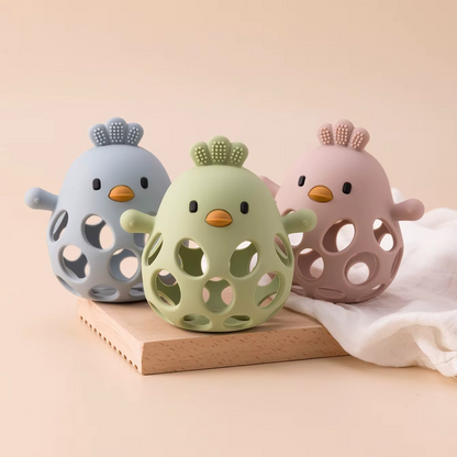 Silicone baby teether rattles in dusty blue, olive green, and rose pink on wooden board with white cloth.