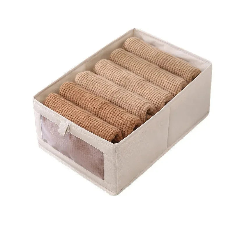 Beige foldable fabric storage box with clear-view front filled with rolled brown baby clothes.