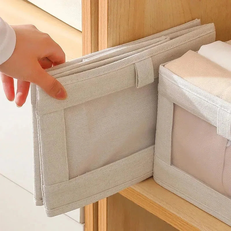 Hand folding empty beige fabric storage box with clear-view front panel beside filled box on wooden shelf.