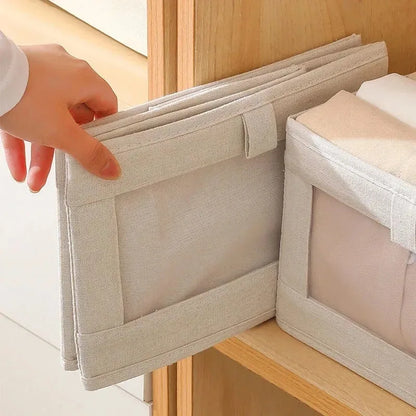 Hand folding empty beige fabric storage box with clear-view front panel beside filled box on wooden shelf.