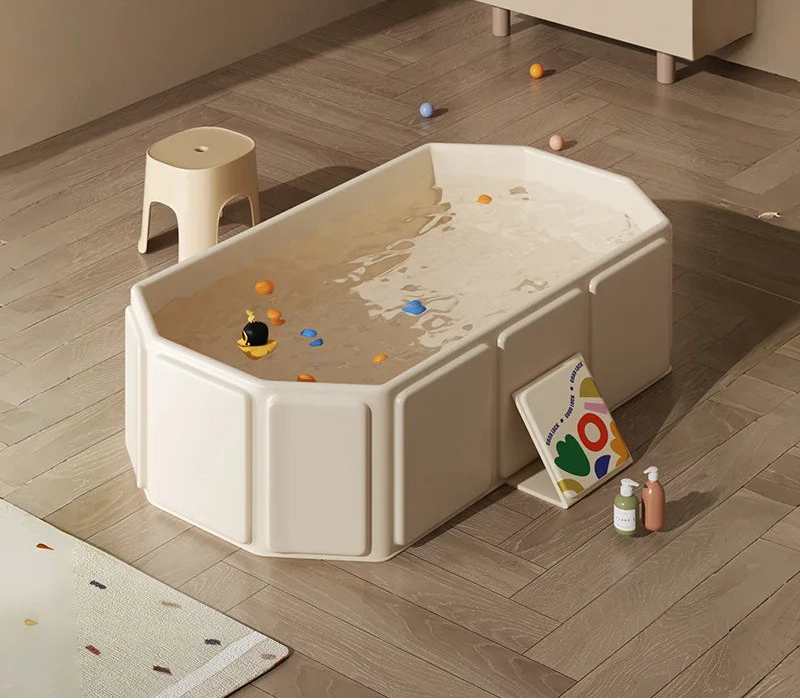 Cream foldable baby bathtub filled with water and colorful balls on wooden floor with bath products.