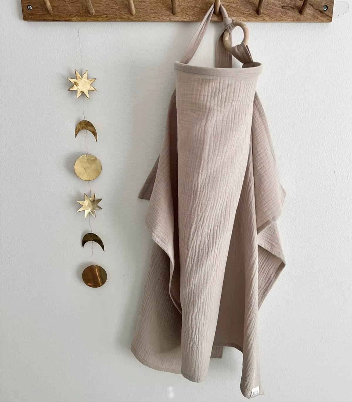 Khaki breathable muslin nursing cover hanging on wall hook with brass sun and moon decor.