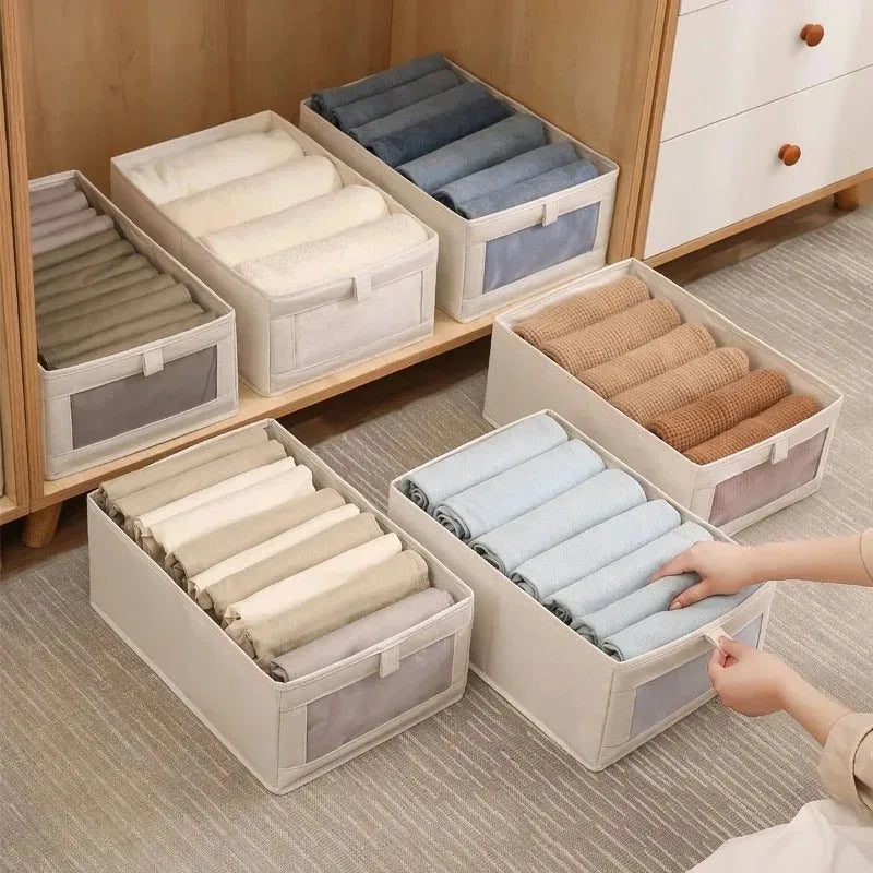 Beige foldable fabric storage boxes with clear-view fronts filled with rolled baby clothes, one box pulled forward by hand.