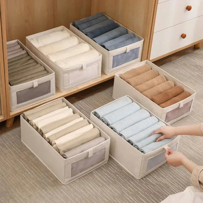 Beige foldable fabric storage boxes with clear-view fronts filled with rolled baby clothes, one box pulled forward by hand.