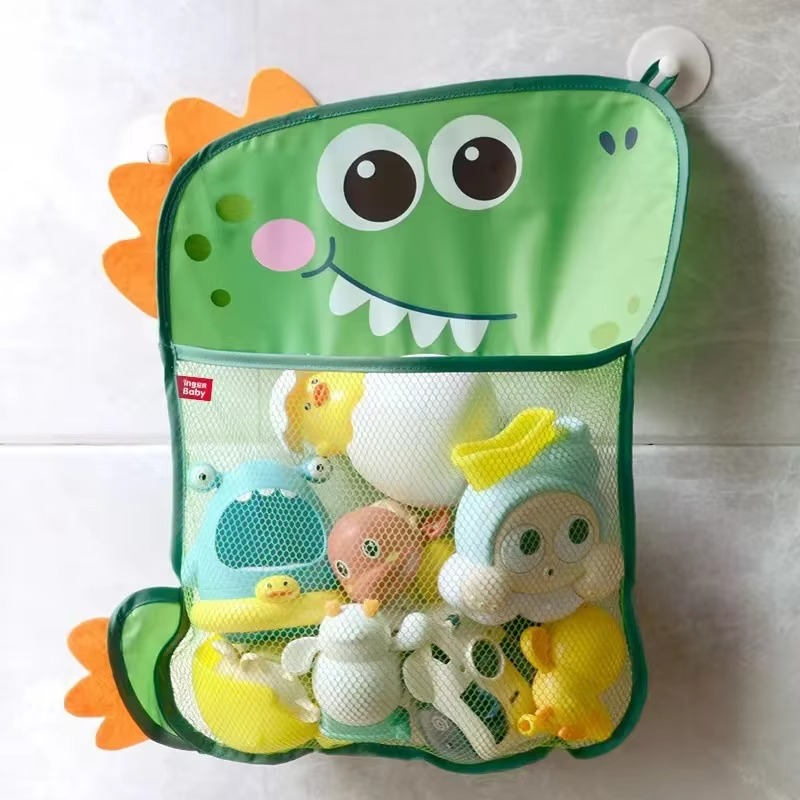 Green dinosaur bath toy storage mesh bag with suction cups holding assorted bath toys on tiled wall.