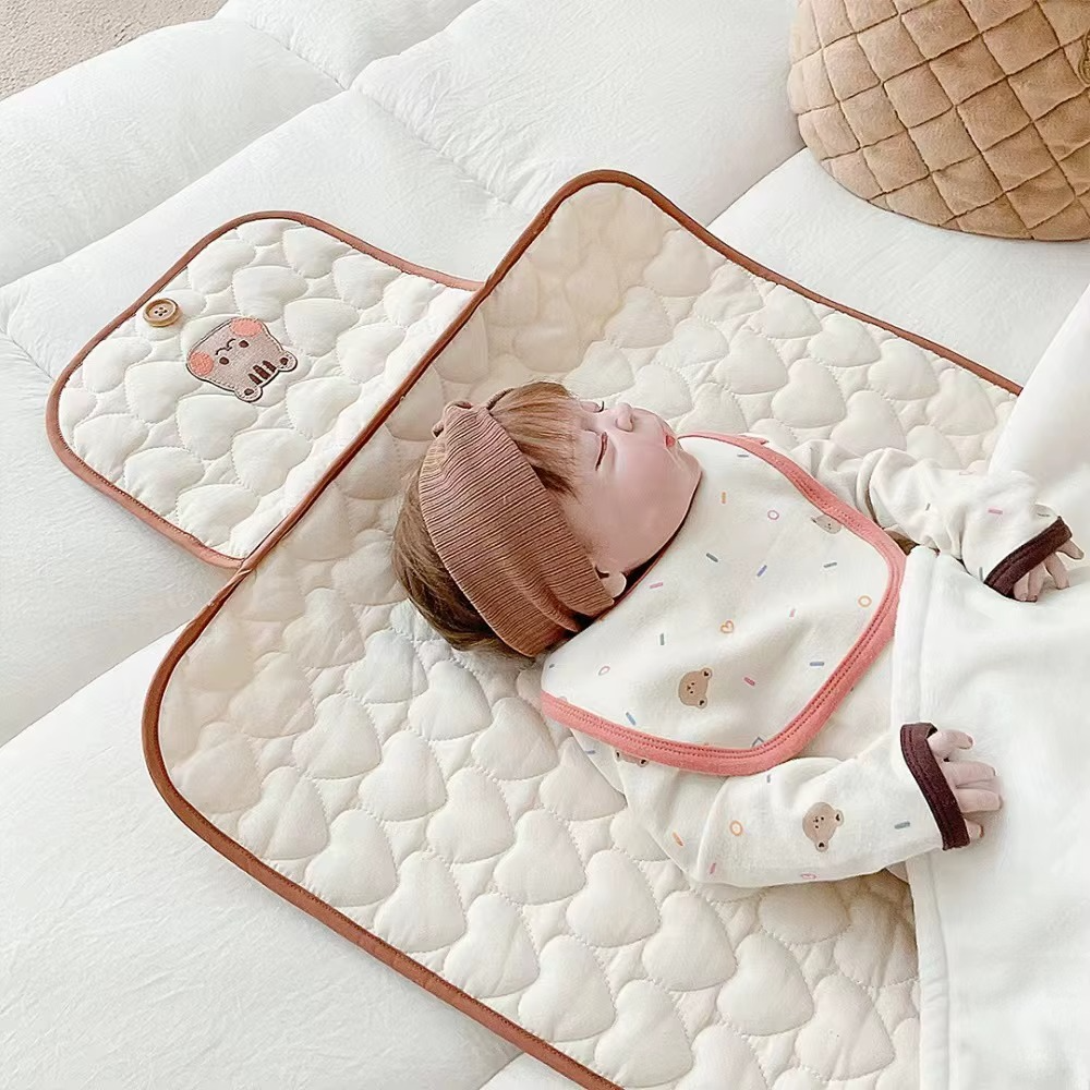Beige foldable quilted baby changing pad with tiger face design in use by baby wearing brown hat.